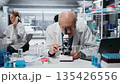 Histology technician analyzing bacterial culture slide to test antibiotic efficacy. Elderly man observing reaction under lens to develop new medical treatments, taking notes, camera B 135426556