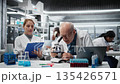 Laboratory technician examining biopsy slide while colleague documents diagnostic findings on clipboard. Elderly man identifies disease markers in clinical lab with coworker taking notes, camera A 135426571