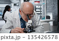 Smiling senior biologist in medical laboratory using scientific microscope for cellular analysis. Jolly elderly man examining sample in lab with high tech optical device gear, camera B 135426578