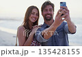 Love, beach and couple taking a selfie with phone enjoying honeymoon, summer vacation and holiday in nature. Travel lifestyle, dating and happy man and woman smile for picture on smartphone by ocean 135428155