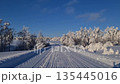 A snow-covered rural road. A snow-white landscape and frost-covered trees along the road. A peaceful snow-white road lined with frost-covered trees in a quiet rural landscape. 135445016