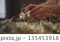 Close-up hands decorating festive garland with glowing house-shaped lights and snowflake details, placing it among tinsel to create warm and cozy holiday atmosphere. Concept of home festive mood. 135453918