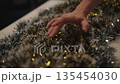 Person hand feeling decorative Christmas tinsel garland with shiny white, green and golden strands, preparing for holiday celebration and winter season traditions, close-up. Shooting in slow motion. 135454030