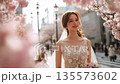 一位身穿白色洋裝的女子，周圍環繞著櫻花花瓣，在櫻花盛開的街道上抬頭望向天空。 135573602