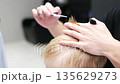 Hairdresser cutting a little blond boy's hair with scissors and comb in a barbershop 135629273