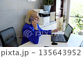 Businesswoman in a blue suit engages in a phone conversation while pointing at her laptop screen, creating a professional and productive office atmosphere 135643952