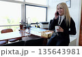 Businesswoman in a tailored suit smiles while conversing on the phone and holding a cup in her sleek office, creating a confident and professional ambiance 135643981