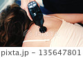 Therapist uses a percussion massage gun on a woman's shoulder while she lies on a treatment table 135647801
