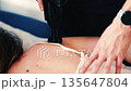 Therapist uses a percussion massage gun on a woman's shoulder while she lies on a treatment table 135647804