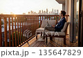 Young businessman working on laptop sits on balcony during golden sunset. Remote laptop work fits labor day career theme or business trip travel lifestyle. Modern city skyline view in background. 135647829