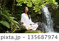 A young Asian woman meditates peacefully on a rock near a waterfall, surrounded by lush green foliage. She wears a white outfit and has long black hair. 135687974