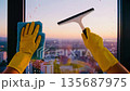A person cleans a large window with a blue cloth and yellow gloves. The city skyline is visible in the background during sunset. 135687975