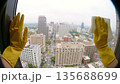 Hands wearing yellow rubber gloves clean a large window overlooking a city skyline. The scene captures urban life and cleanliness. 135688699