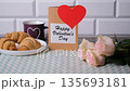 A male hand is placing a postcard with a greeting and a gift box on a table. The table has breakfast items like croissants, juice, tea, and a bouquet of flowers 135693181
