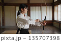 A female high school student in the archery club slowly draws her bow and aims at the target in the dojo. A female high school student in the archery club slowly draws her bow and aims at the target in the dojo. 135707398