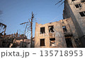 urban destruction, bombing aftermath, war ruins. Urban destruction scene showing severely damaged buildings, broken windows, charred trees under clear blue sky after recent bombing. 135718953