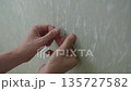 Close-up hands of man removing old transparent double-sided adhesive tape from wall, showing effort of cleaning and preparing surface for renovation or redecoration. Shooting in slow motion. 135727582