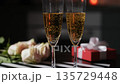 Bubbles rise in glasses of champagne on a table. There is a bouquet of roses and a gift box nearby. This captures a cozy atmosphere for Valentines Day celebration at home with loved ones 135729448
