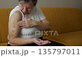 Old Woman Massages, Elderly Woman Soothing Arm Injuries Calmly, Elderly Woman Gently Pressing Bruised Arm In Peaceful Room, Senior Woman Tenderly Rubbing Her Injured Arm On Cozy Yellow Couch At Home 135797011