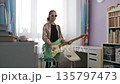 A girl in sunglasses plays electric guitar at home. She explores her passion for music and enjoys the creative process. She stands in a colorful room filled with her musical items and inspiration 135797473