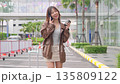 Female traveler enjoys beautiful city holidays, woman along modern street, tourist talking on phone, holding coffee, pulling luggage, urban architecture surrounds. 135809122