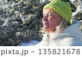 Senior woman with eyes closed breathing in fresh frosty air in snowy winter park on sunny day. Female enjoying serene moment and warmth of sun on face. Sunbathing to produce vitamin D. 135821678