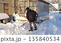 Person shovels cold winter snow. Individual removes heavy snow in rural area. Man working outdoors to clear thick winter snow drifts. Person diligently shovels snow outside shed in cold rural morning 135840534