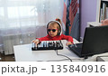 A little girl enjoys crafting music on a midi keyboard at home. She wears headphones and sunglasses while shaking her head to the rhythm, showing her love for music 135840916