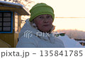 Mature caucasian woman with blond hair wearing warm winter clothes. Adult female looking at camera while standing outside in snowy village during beautiful cold and sunny sunset. Breath vapor in cold 135841785