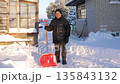 Elderly man in warm winter clothes standing in backyard with snow shovel. Looking at camera after clearing path from the heavy snowfall during sunny cold day. Person clearing snow outdoors. 135843132
