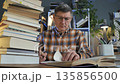 Man studying book at desk with large stack of books in home interior 135856500