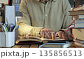 Hands reading open book at desk surrounded by stacked books indoors 135856513
