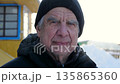 Close up portrait of elderly man with gray hair and blue eyes. Old man dressed in winter hat and down jacket looking at camera on clear snowy day in courtyard of house. Old face with deep wrinkles 135865360
