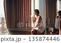 Playful young woman wrapped in a towel blows foam from her hands while enjoying a relaxing morning 135874446