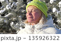 Content older woman with closed eyes enjoying sunshine and fresh air in winter forest. Wearing warm jacket and green knitted hat while standing against background of snowy pine trees 135926322
