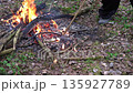 Amidst the serene woodland floor, a person carefully stokes a vibrant campfire, shifting burning branches with a stick 135927789