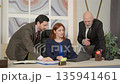 Confident charming redhead mature female CEO and handsome male colleagues gathering around laptop, discussing and sharing ideas about business project while working at office. 135941461