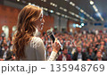 Young female speaker in crowded business audience gives speech. Woman is on commercial conference session. Lecture in exposition hall 135948769