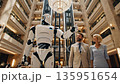 Tilt-up video of a humanoid robot guiding a couple through a hotel's front desk and carrying their luggage 135951654
