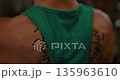A close-up captures muscular man's back featuring tattoo, wearing green tank top in gym. This showcases fitness, dedication, and healthy lifestyle, highlighting strength and wellness. 135963610