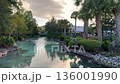 A narrow canal of clear turquoise water winds between lush green trees and palms. Rocks and landscaped bushes create a peaceful tropical park scene at sunset. 136001990