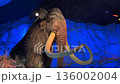 A towering mammoth statue with long ivory tusks rises against glowing blue ice walls. Warm spotlights highlight the animal's fur inside the themed prehistoric cavern. 136002004