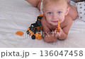 A little boy of one year old with blond hair and blue eyes lies on a bed and chews a toy looking at the camera. High quality 4k footage 136047458