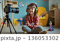 A little girl sitting on the floor with headphones on and a camera 136053302