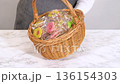 A person dressed in a neutral apron holds a wicker basket filled with colorful flower-shaped cookies wrapped in plastic. The basket is shown steady at waist level, conveying a sense of care and gift 136154303