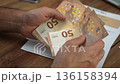 Male office worker meticulously transfers stack of 50-euro banknotes from hand to other counting money, man puts hands on wooden table with documents referring to accounts 136158394
