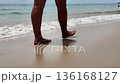 A close-up of a woman's legs strolling along the seashore on a sunny day, with gentle waves lapping at the sand in the background. Malaysia 136168127