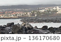 Wide view of waves breaking on dark rocks along a coastal resort town, palm trees and buildings in the distance, moody overcast sky and sea spray. Ideal for travel, weather, and nature concepts. 136170680