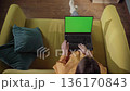Person relaxing on a yellow sofa with feet up, using a laptop with a chroma key green screenperfect mockup for remote work, online learning, video calls, or leisure at home 136170843