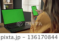 Woman using a smartphone with a green screen while a laptop with a green screen sits on a wooden desk, representing remote work, digital communication, and online education concepts 136170844